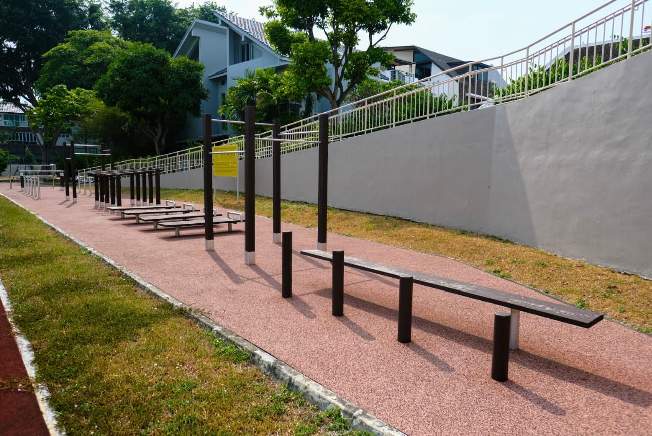 Outdoor fitness area with benches and pull-up bars on a red rubber path beside a gray wall and green trees.